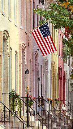 an american flag flying in the wind on a city street lined with multi - colored buildings