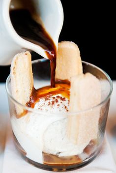 someone pouring caramel syrup into a small glass bowl filled with ice cream and marshmallows