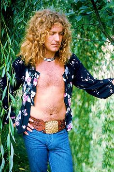 a man with long hair and no shirt is standing in front of some trees wearing blue jeans