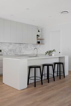 a white kitchen with marble counter tops and stools next to an island that has three plants on it