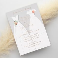 a wedding card with a dress and flowers on it next to some pamy feathers