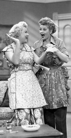 two women in aprons are talking to each other while standing near a coffee table