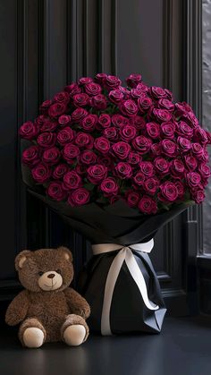 a brown teddy bear sitting next to a large bouquet of purple roses in a black vase