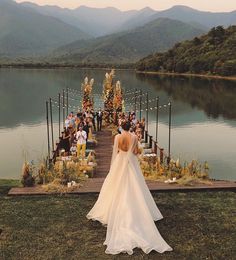 a woman in a wedding dress is standing on the edge of a dock with mountains in the background