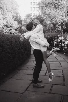 a man and woman kissing in the middle of a walkway surrounded by bushes with flowers