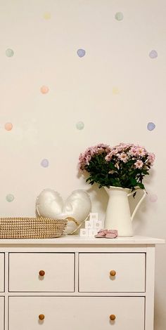 a white dresser topped with a vase filled with pink and purple flowers next to a wall covered in polka dot decals