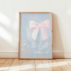 a painting with the words be the light and a pink bow on it in front of a white wall