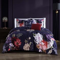 a bed covered in purple and red floral comforter sets with pillows on top of it