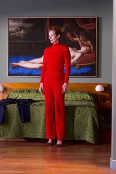 a woman standing in front of a bed wearing a red jumpsuit and matching heels