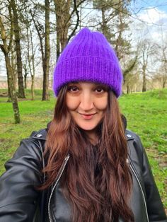 a woman wearing a purple knitted hat and black leather jacket standing in front of trees