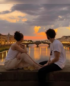 two people are sitting on a ledge overlooking the water and bridge at sunset or dawn