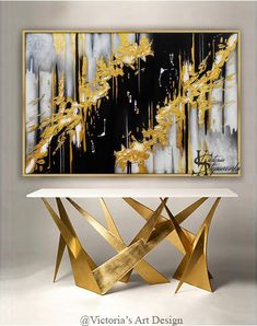 an abstract painting with gold and black accents on a white table in front of a gray wall