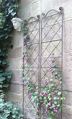 an iron garden trellis with purple flowers growing on it's sides, next to a brick wall