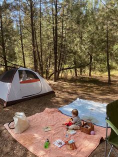 a little boy sitting in front of a tent on top of a blanket next to a picnic table