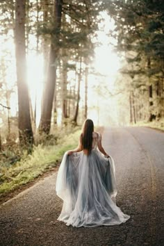 a woman is walking down the road with her dress blowing in the wind and sun shining through the trees