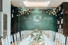 a long table with white chairs in front of a green wall and floral decorations on it