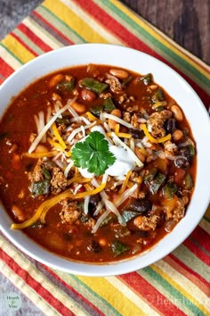 a white bowl filled with chili and beans on top of a colorful table cloth next to a spoon