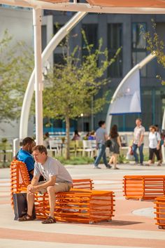 two men sitting on orange park benches in front of a building with people walking by