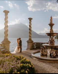 a bride and groom standing in front of a fountain with mountains in the back ground