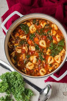 a pot filled with pasta and spinach soup on top of a red cloth next to a spoon