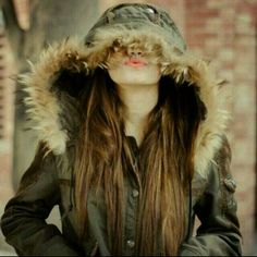 a woman with long hair wearing a jacket and hood on her head is standing in front of a brick building