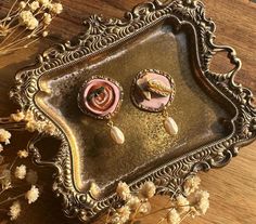 two pink and gold earrings sitting on top of a metal tray next to dried flowers