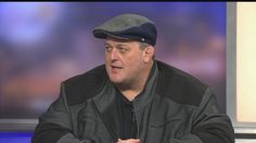 a man in a black jacket and hat is talking on the set of an interview