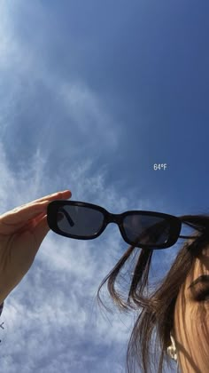 a woman is holding up her sunglasses to the sky with clouds in the back ground