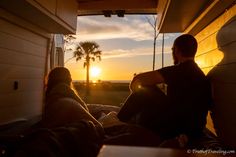 two people sitting in the back of a truck watching the sun go down with palm trees behind them