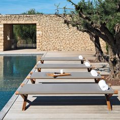 several lounge chairs sitting next to a pool in front of a stone wall and tree