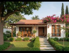 a small white house with lots of flowers on the roof and bushes around the front door