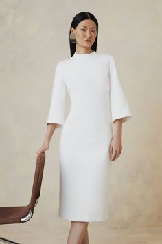 a woman standing next to a chair wearing a white dress with bell sleeves and heels