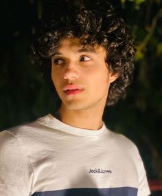 a young man with curly hair wearing a white t - shirt looking at the camera