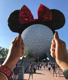 a person holding up a minnie mouse head in front of a disney world building with people walking around it