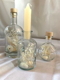 three glass bottles with flowers in them sitting on a white cloth covered tablecloth next to a lit candle