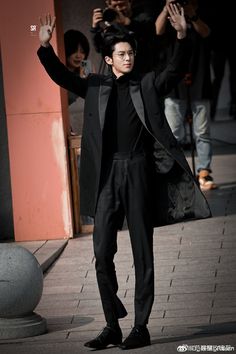 a man in black suit and tie walking down the street with his hands up to him