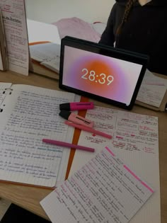 a desk with notebooks, pens and a computer screen displaying the date on it