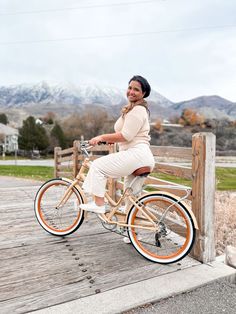 a woman sitting on top of a bike next to a wooden fence with mountains in the background