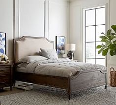 a bedroom with a bed, nightstands and plants on the floor in front of large windows