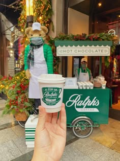 a person holding up a cup of coffee in front of a hot chocolate stand at christmas time