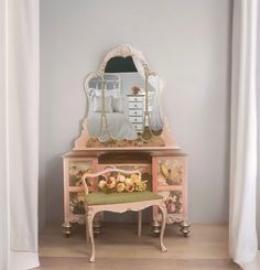 a pink dresser with a mirror and flowers on it in front of a white curtain