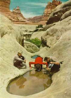 three people sitting on rocks in the desert drinking water from a small pond that is surrounded by large rock formations