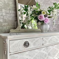 a white dresser with flowers on top and a mirror above it in front of a brick wall