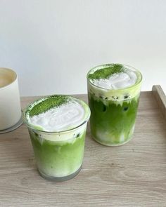 two glasses filled with green drinks sitting on top of a table next to a cup