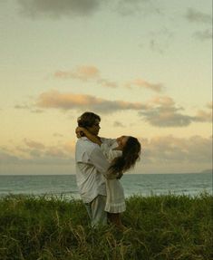 a man and woman are standing in the grass by the ocean at sunset or sunrise