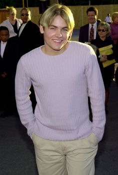 a young man in a sweater and khaki pants smiles at the camera as people look on