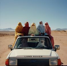 three women in burkas sitting on the hood of a car with desert background