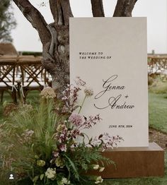 a wedding sign sitting in the grass next to a tree and flower potted plant