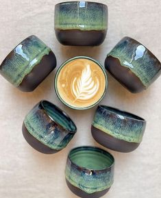 there are six cups with different designs on them and one has a latte in the middle