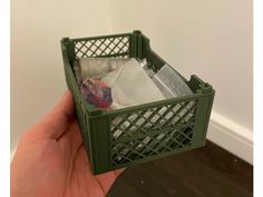 a hand holding a green plastic container filled with garbage next to a white wall and wooden floor